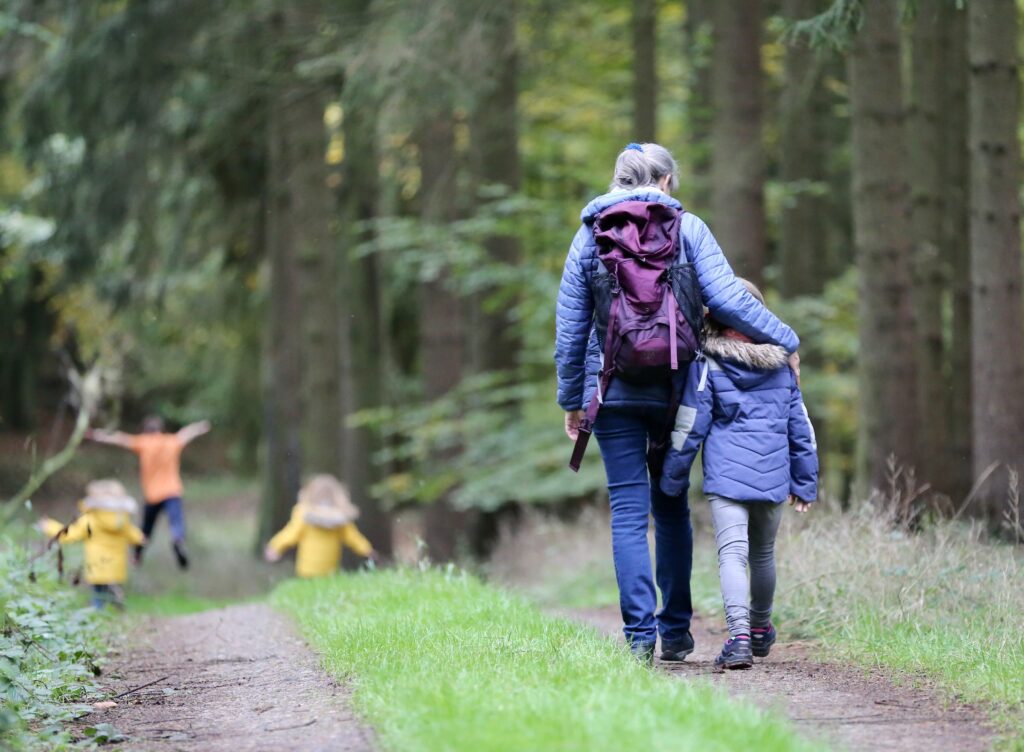 woman in blue denim jeans and blue jacket walking with child in blue jacket through a forest. Three other children in yellow play in the distance